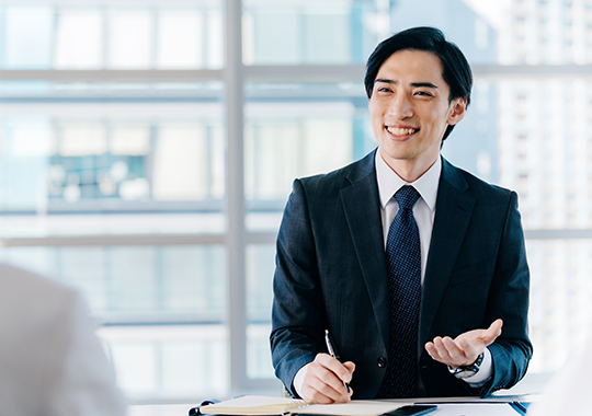 経営コンサルサービスのイメージ - プロフェッショナルな男性がオフィスでデスクに座り、ペンを持って笑顔で対応している様子